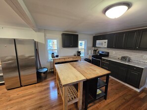 Une cuisine avec un plancher en bois, un réfrigérateur en acier inoxydable, une table en bois, des meubles de couleur sombre, un four à micro-ondes et une cuisinière.