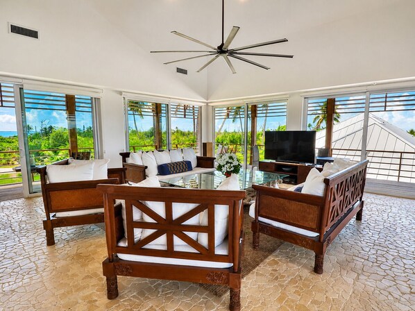 A spacious living area with a glass coffee table, wooden furniture, a television, and large windows offering a view of the outdoors.