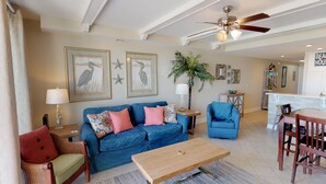 A living room with a blue sofa, a wooden coffee table, a wicker chair, and a blue armchair. There are framed wall art, a ceiling fan, and a kitchen area in the background. - Beachfront Luxury 1 BR Condo - 3 Oceanfront Pools (Tybee Island)