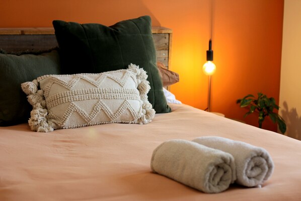 A neatly made bed with a white knitted blanket and rolled towels, a wooden headboard, and a bedside lamp.