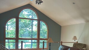 Un vaste salon doté d’un haut plafond, d’un ventilateur de plafond, d’un canapé en cuir marron, d’une table basse en bois et de grandes fenêtres en arc de cercle offrant une vue sur les arbres.