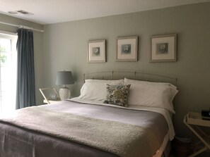 Un lit bien fait, avec une couette blanche et des coussins à motifs floraux, une table de chevet avec une lampe, et trois tableaux encadrés accrochés au mur.