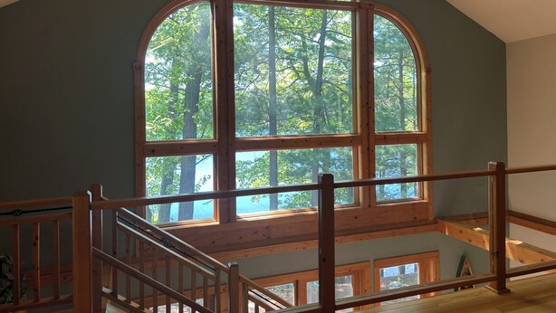 Une pièce dotée d’une grande fenêtre en arc d’où l’on aperçoit une forêt, d’un escalier en bois et d’une vue sur le ciel.