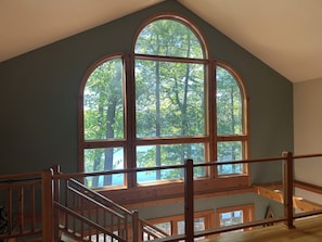 Une pièce dotée d’une grande fenêtre en arc d’où l’on aperçoit une forêt, d’un escalier en bois et d’une vue sur le ciel.