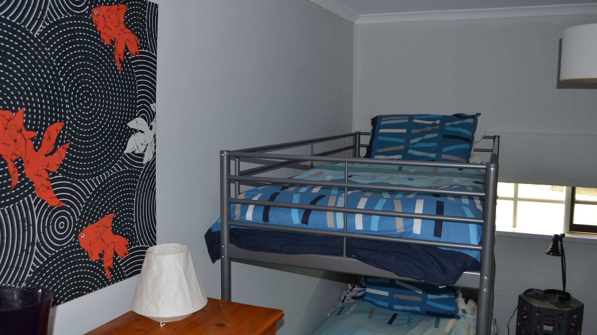 A bunk bed with a blue and white striped bedspread, a white lamp on a wooden table, and a wall with a black and red abstract design.