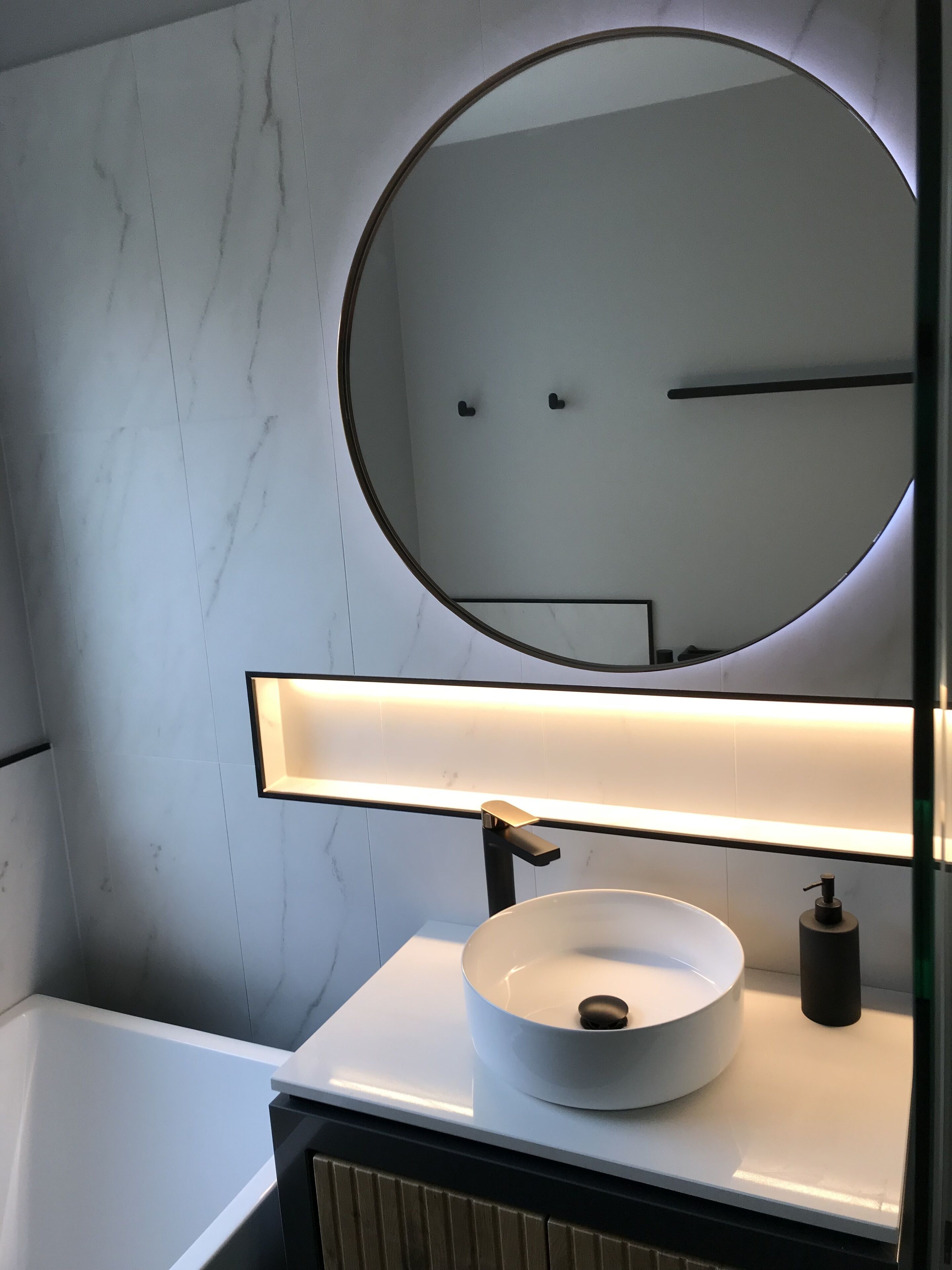 Une salle de bain avec un miroir rond, un lavabo blanc et un luminaire fixé au mur.