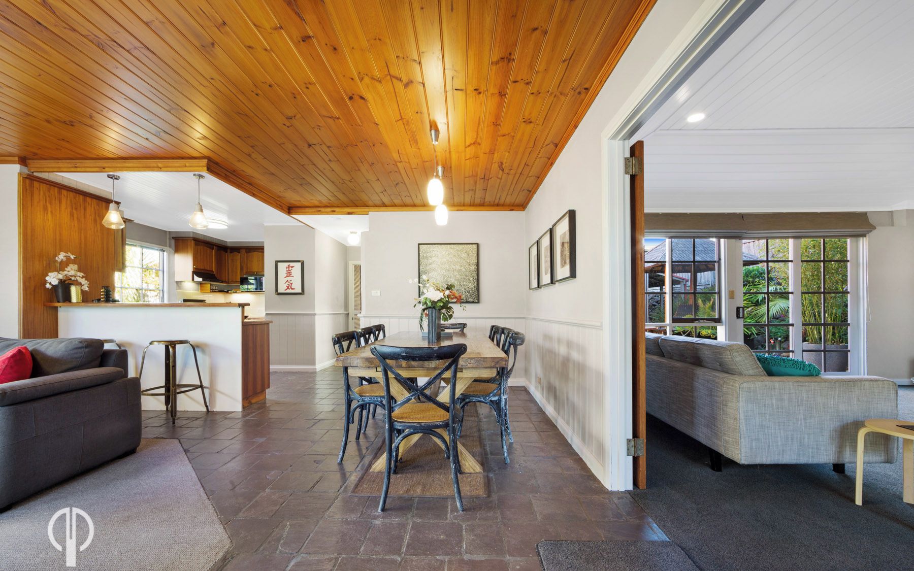 A spacious living area with a wooden ceiling, a dining table with chairs, a sofa, and a kitchen in the background.