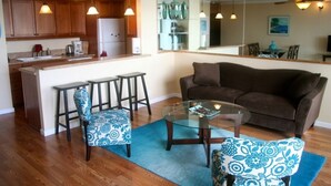 A modern kitchen with wooden cabinets, a refrigerator, and bar stools. There is a glass coffee table and a brown sofa in the living area. - Unobstructed ocean view 2Br/2Ba - upscale at a budget price (Kihei)