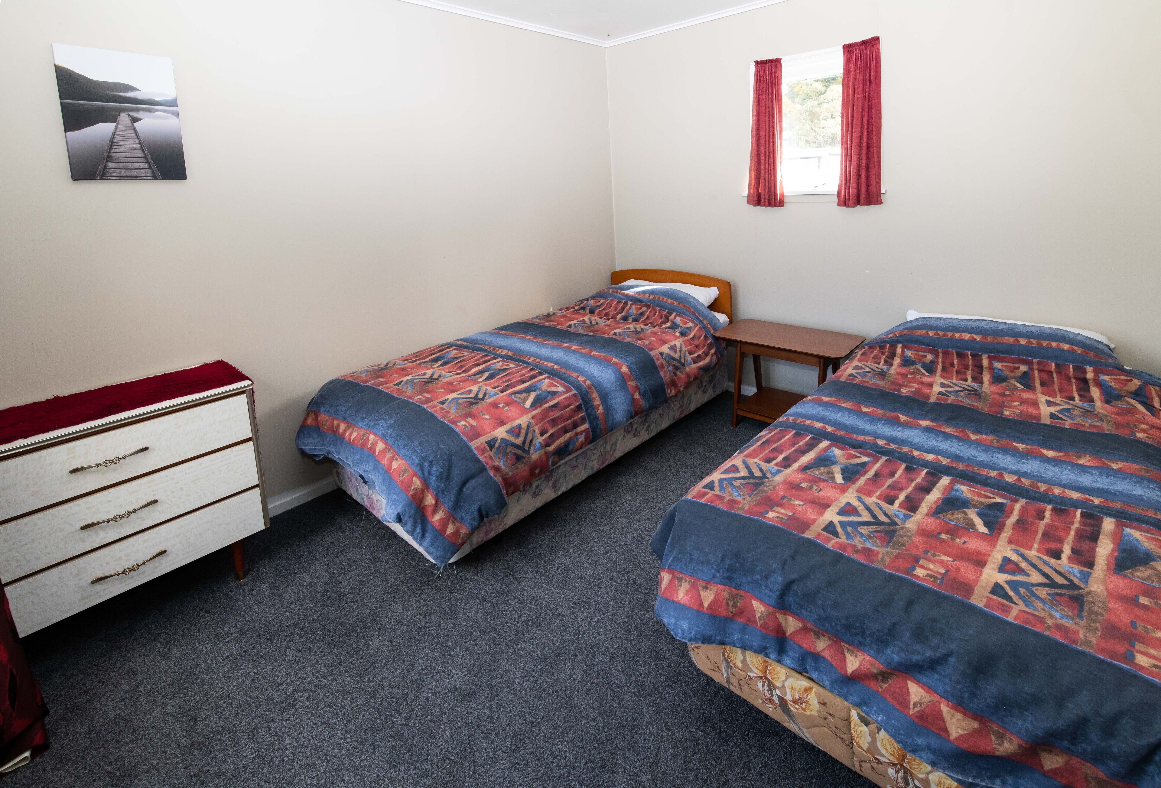Une chambre avec deux lits simples, une commode en bois, une petite table et une fenêtre avec des rideaux rouges.