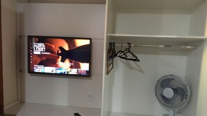 Un televisor montado en la pared mostrando una escena de película, un control remoto en un estante y un ventilador de pie.