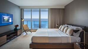 A modern bedroom with a large bed, a TV displaying a golf channel, a chair, and a view of the ocean. - Incredible Ocean Views! 2 BR/2.5 Bath, On Top of 1 Hotel! 15th Flr, 4 Pools! (Miami Beach)