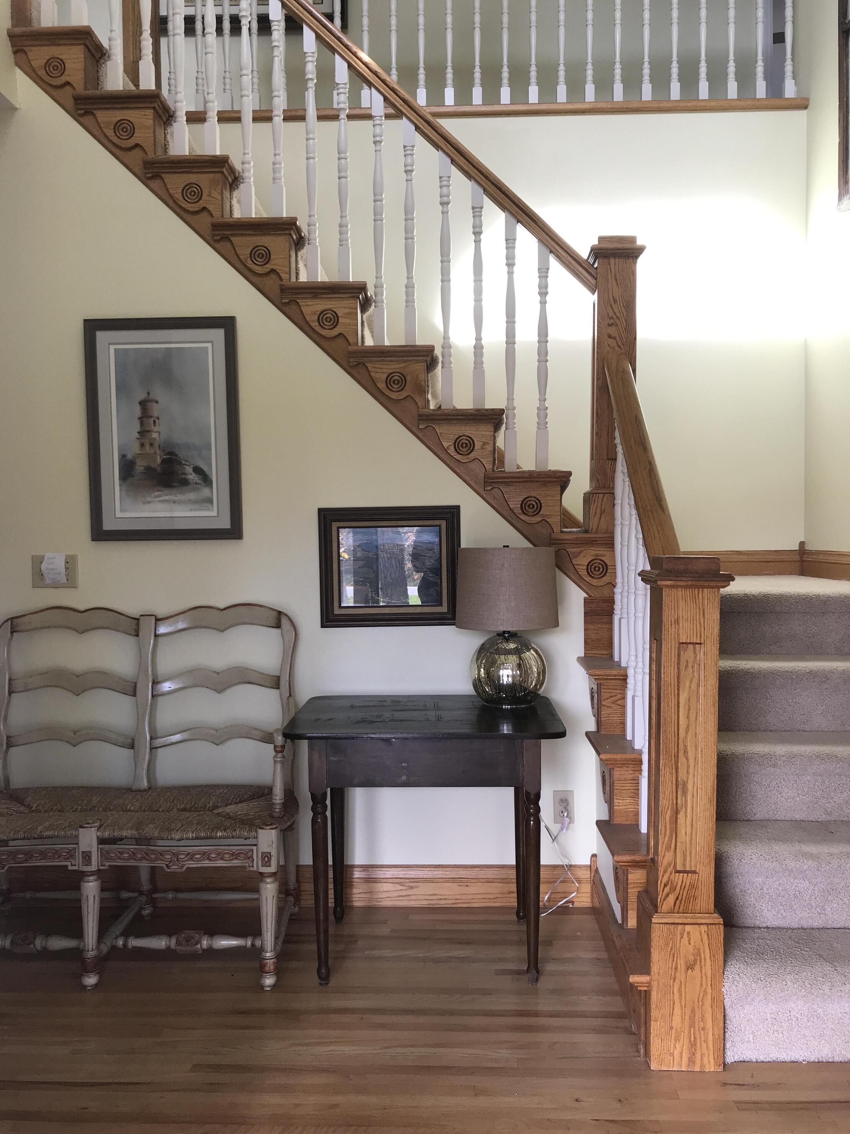 Una escalera de madera con balaustres blancos, un cuadro enmarcado de una torre, una mesa con una lámpara y un banco.