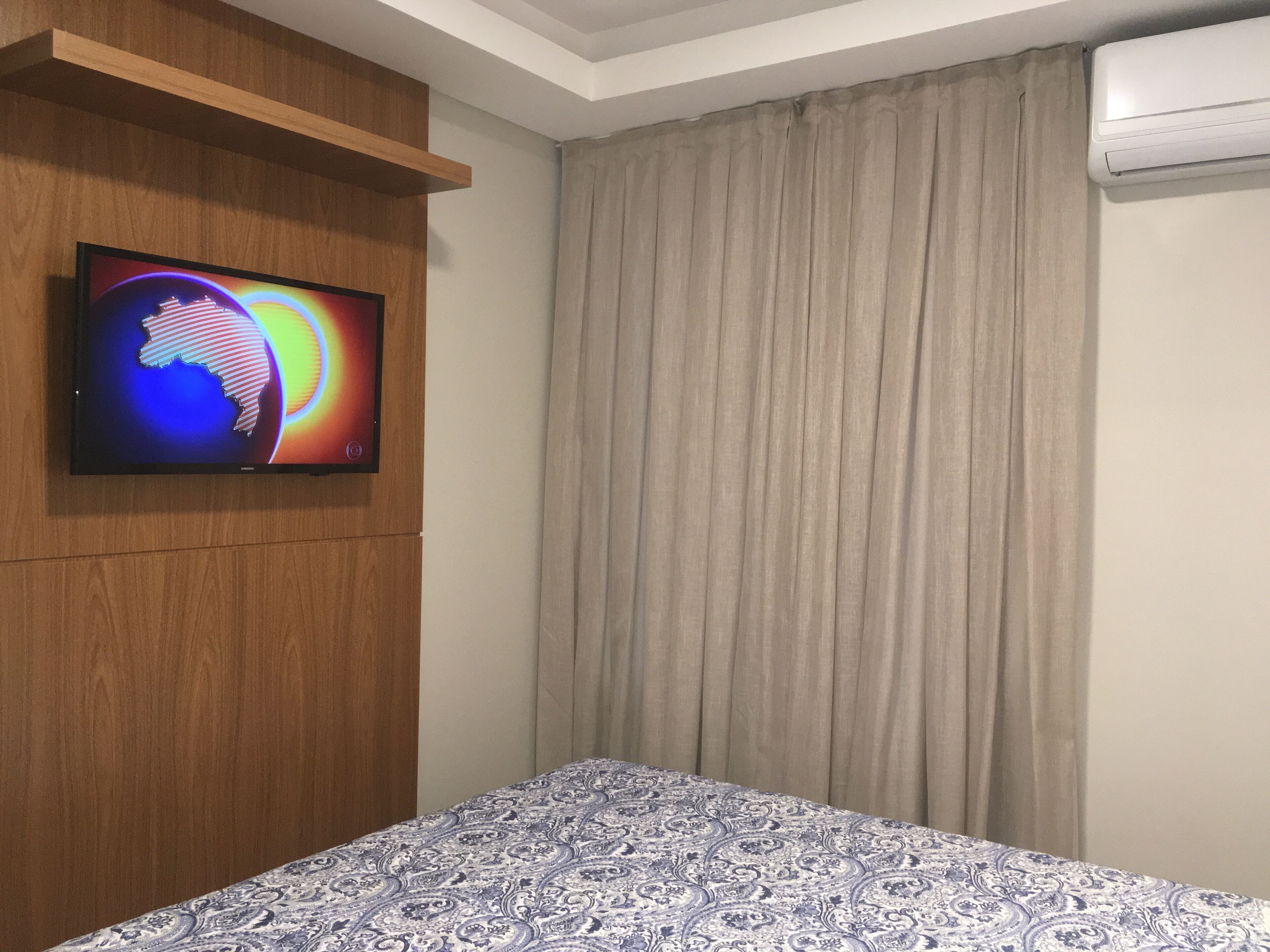 A hotel room with a bed, a television mounted on a wooden panel, a window with beige curtains, and an air conditioning unit.