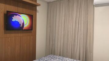 A hotel room with a bed, a television mounted on a wooden panel, a window with beige curtains, and an air conditioning unit.