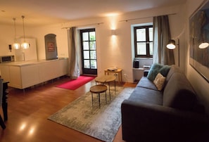 A living room with a sofa, a coffee table, a rug, a kitchen area with a microwave, and a window with curtains. - Newly Renovated! Perfect home away in Munich. (Munich)