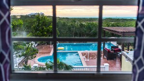 A view of a swimming pool with a wooden deck and lounge area, surrounded by greenery and a distant view of a house.