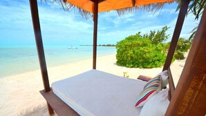 A bed with a white mattress and colorful pillows, set against a beach backdrop with clear blue water and a thatched roof structure.