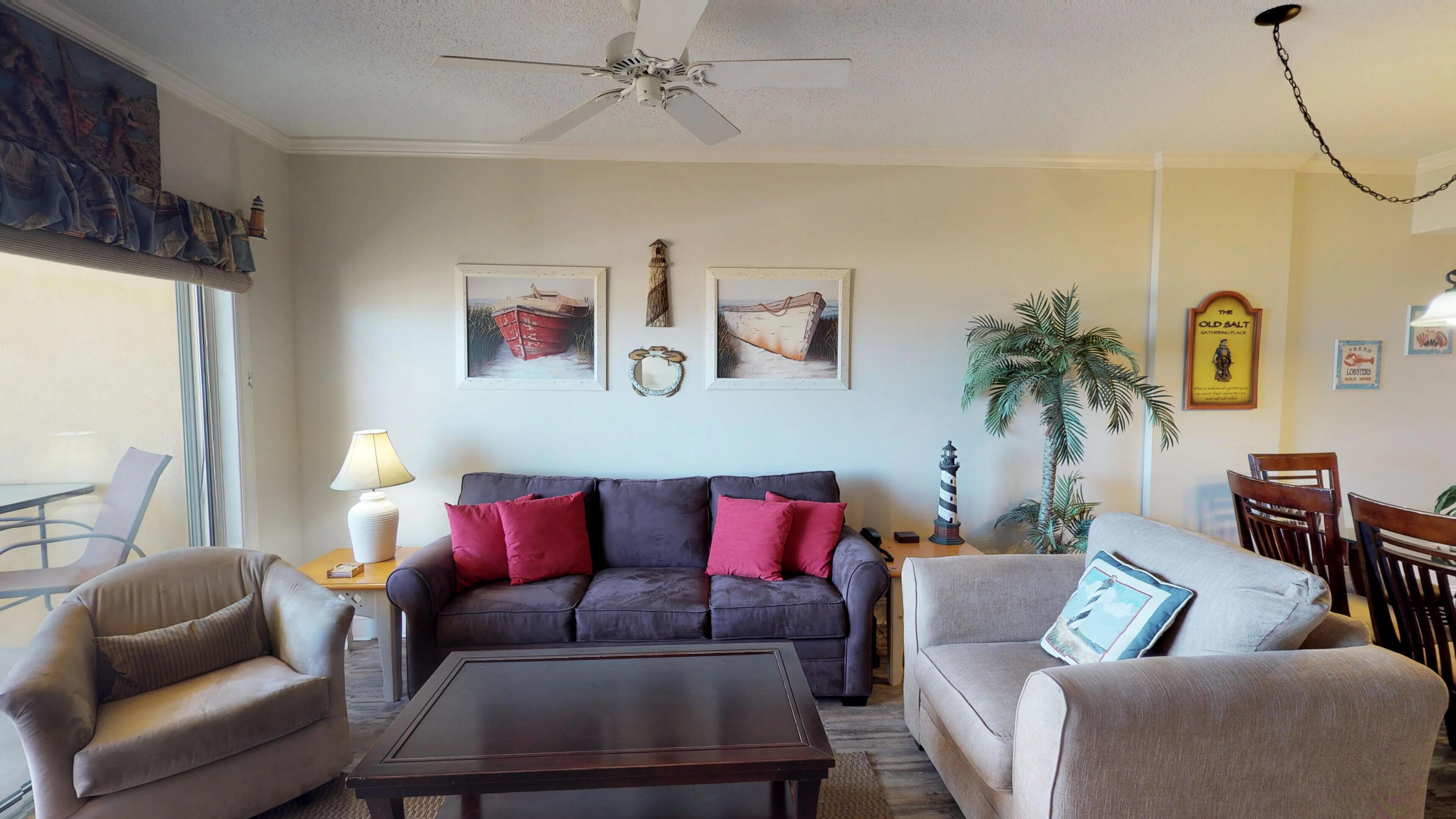 A living room with a dark-colored sofa, a beige armchair, and a wooden coffee table. There is a ceiling fan, a lamp, and framed artwork on the wall.