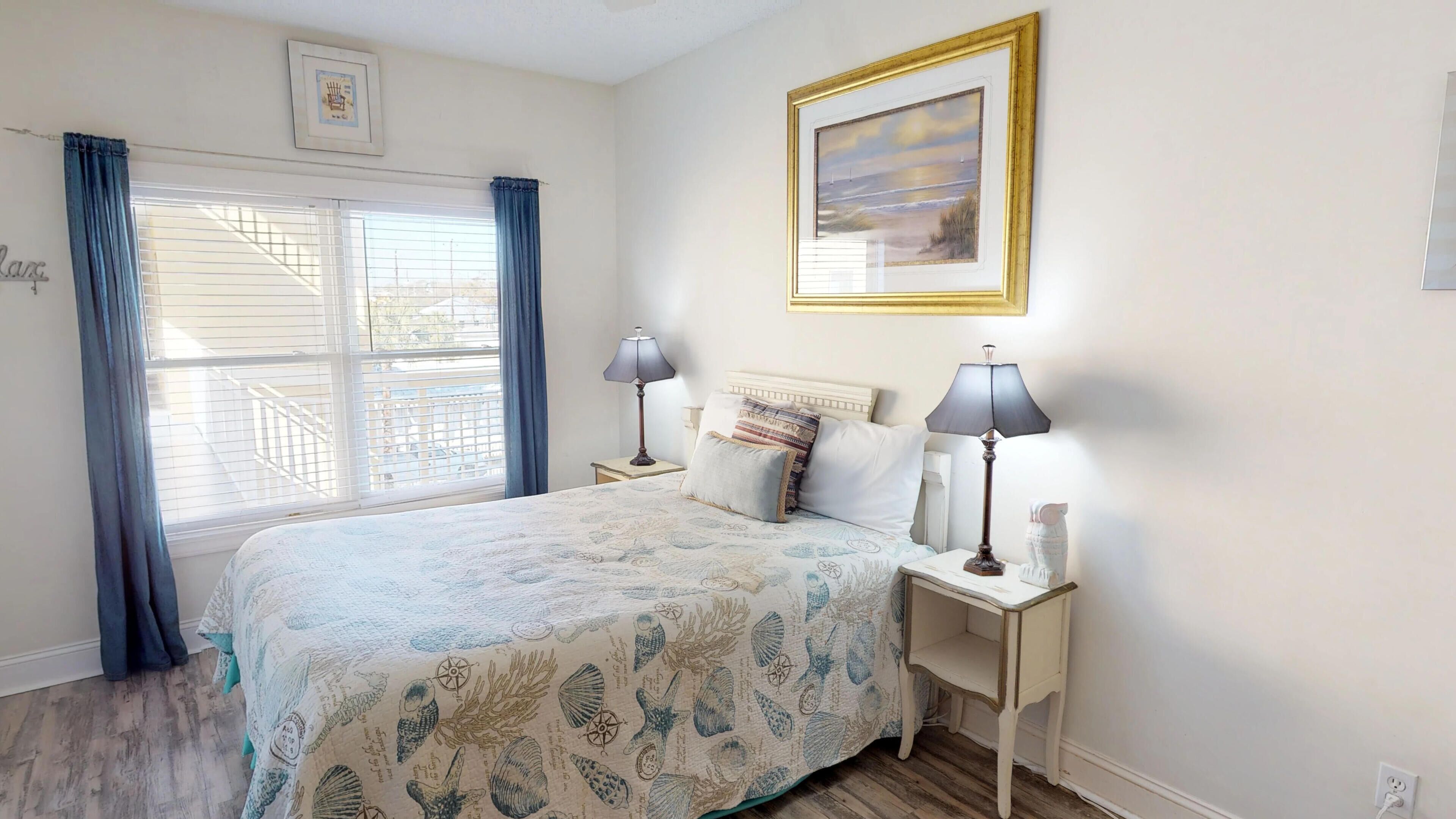 A bedroom with a bed, two bedside tables with lamps, a window with curtains, a framed picture on the wall, and a small shelf with decorative items.