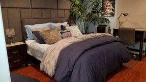 Um quarto com cama, mesas de cabeceira, escrivaninha, cadeira, abajur e uma planta.