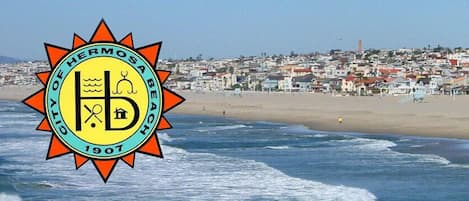 Una playa con el logo de Hermosa Beach, 1907, y un pueblo costero al fondo.