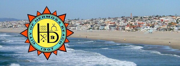 Una playa con el logo de Hermosa Beach, 1907, y un pueblo costero al fondo.