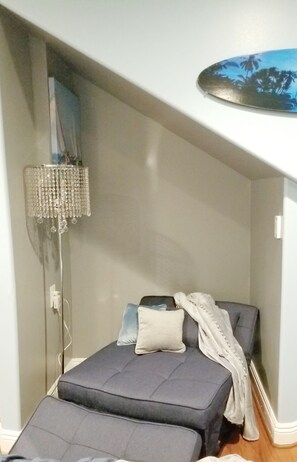 Quarto pequeno e compacto com cama, mesa de cabeceira, abajur e prateleira na parede.