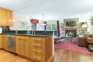 A modern kitchen with wooden cabinets and a central island, a living area with a fireplace, and a dining space with a red rug. - 100 ft. to Gondola!! Balcony, Fireplace, Views, Parking!! Heart of Lionshead!! (Vail)