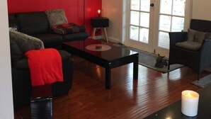 A living room with a red wall, a black sofa, a coffee table, and a window with blinds. - Modern Cottage in peaceful setting, dog door to large fenced yard, farm walks.  (Mil-lel)