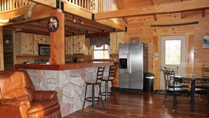 A wooden cabin interior with a kitchen, a stone accent wall, a leather sofa, and a dining area with metal chairs.
