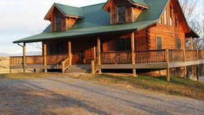 A log cabin with a green metal roof, a porch with wooden railings, and a gravel driveway.