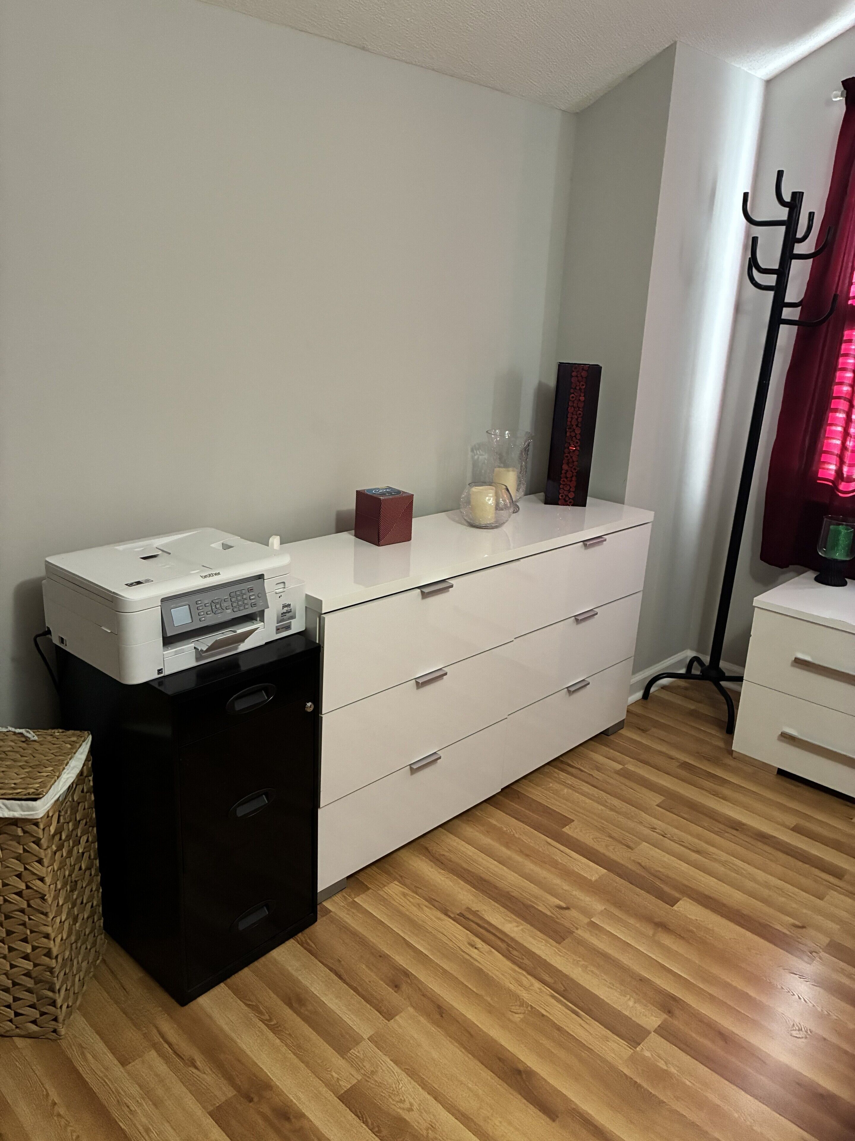 Une pièce comprenant une imprimante posée sur un support noir, une commode blanche avec des tiroirs, un panier en osier et une lampe sur pied.
