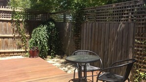 Eine Holzterrasse mit rundem Tisch und zwei Stühlen, umgeben von einem Holzzaun und Grünpflanzen.