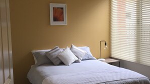 A bedroom with a bed, pillows, a bedside table with a lamp, a framed picture on the wall, and a window with blinds. - Healesville Cottage - 2 Bedroom House (Healesville)