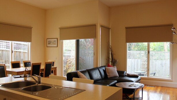 A spacious living area with a black leather sofa, a dining table with chairs, a kitchen with a double sink, and large windows offering views of a backyard with a wooden fence. - Healesville Cottage - 2 Bedroom House (Healesville)