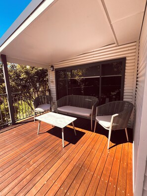 Une terrasse extérieure couverte, dotée d’un plancher en bois, de deux fauteuils en osier, d’une petite table et d’une grande fenêtre.