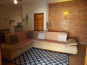 Un salon avec un canapé d’angle, un tapis à motifs, un mur en briques et une porte en bois.