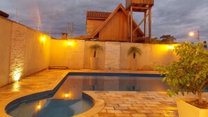A swimming pool area with a wooden deck, potted plants, and a building with a sloped roof.