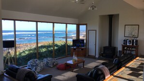 A spacious living room with a large window overlooking a beach, a television, a fireplace, and a bookshelf. - Bluestone Bay - Port Fairy (Port Fairy)