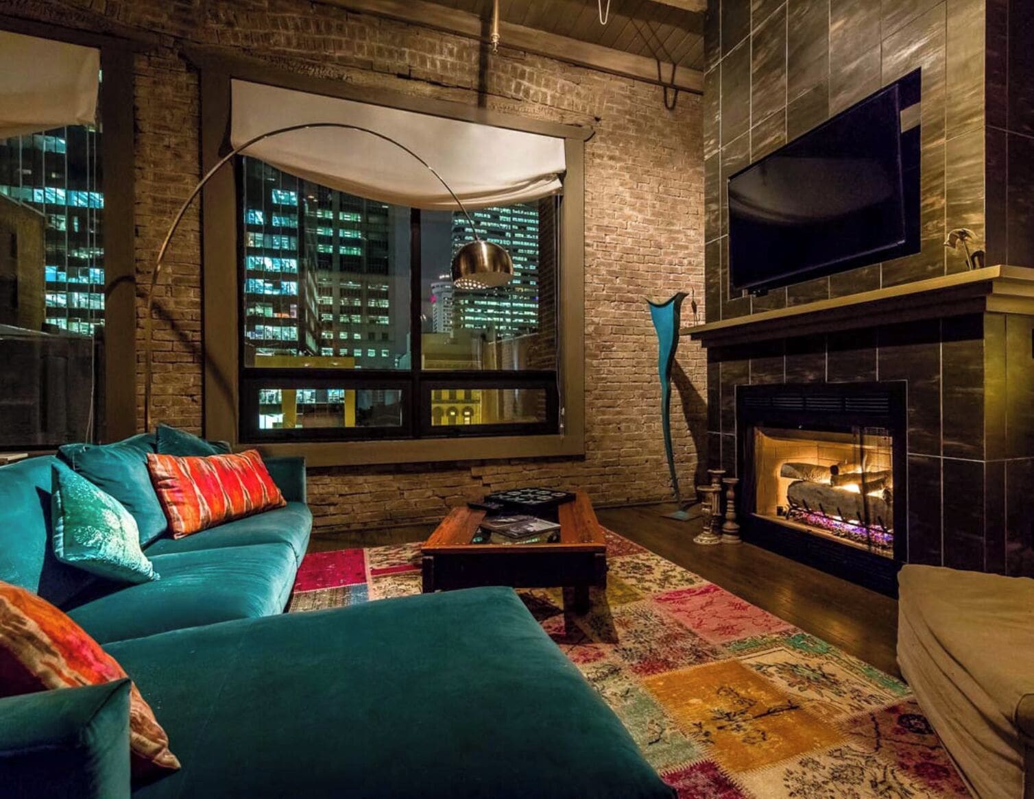 A cozy living room with a brick wall, a large window overlooking a cityscape, a fireplace, and a flat-screen TV mounted above it.