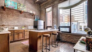 A kitchen with a brick wall, wooden cabinets, a central island, bar stools, and a window with a city view. - City Loft PH*Walk to Broadway*Printers Alley Lofts (Nashville)