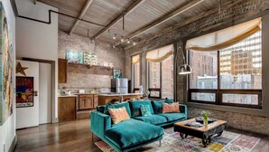 A spacious living room with a teal sofa, a coffee table, and a colorful rug. There is a kitchen area with a brick wall and a window overlooking the city. - City Loft PH*Walk to Broadway*Printers Alley Lofts (Nashville)
