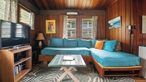 Ein gemütliches Zimmer mit Holzbalkendecke, einem großen Flachbildfernseher, einem Sofa mit blauer Bettwäsche und einem kleinen Tisch mit einem Buch.
