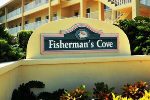 A sign for Fisherman's Cove with a decorative shell emblem and a balcony in the background.