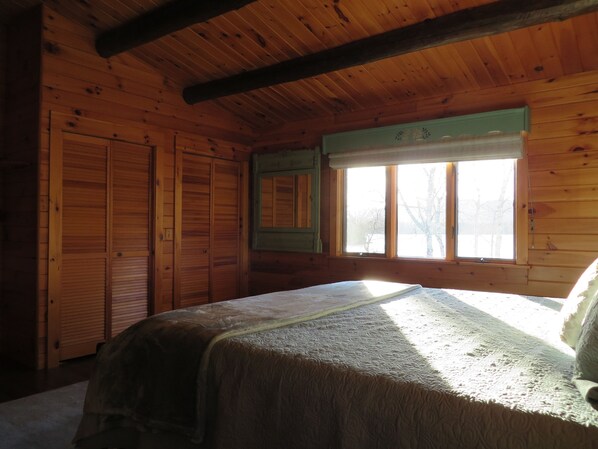 A wooden cabin room with a bed, a window with a view of trees, and a green-framed mirror.