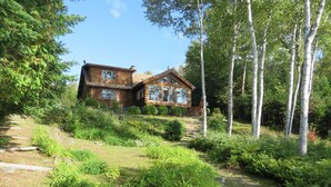 A rustic wooden cabin with a stone pathway leading to it, surrounded by lush greenery and birch trees. - SUNDOWN ON MOOSEHEAD LAKE - PLEASE READ THE ENTIRE DESCRIPTION  BEFORE BOOKING. (Greenville)