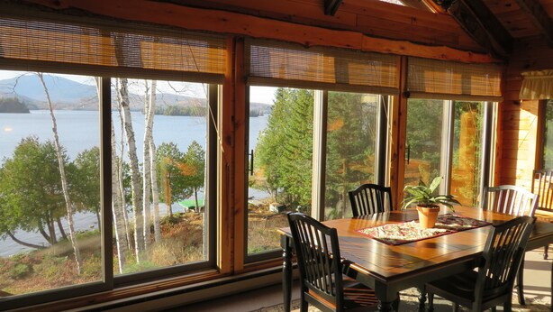 A dining area with a wooden table and chairs, large windows offering a view of a lake and trees. - SUNDOWN ON MOOSEHEAD LAKE - PLEASE READ THE ENTIRE DESCRIPTION  BEFORE BOOKING. (Greenville)