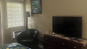 Une chambre à coucher comprenant un lit, un téléviseur, une chaise, une commode, un ventilateur de plafond et une fenêtre avec des stores.