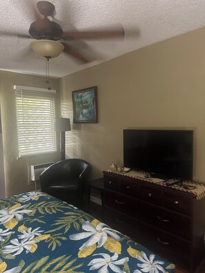 Un dormitorio con cama, televisión, sillón, cómoda, ventilador de techo y ventana con persianas.