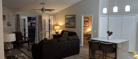 A modern living room with a dark sofa, a dining area with bar stools, a ceiling fan, and a painting on the wall.